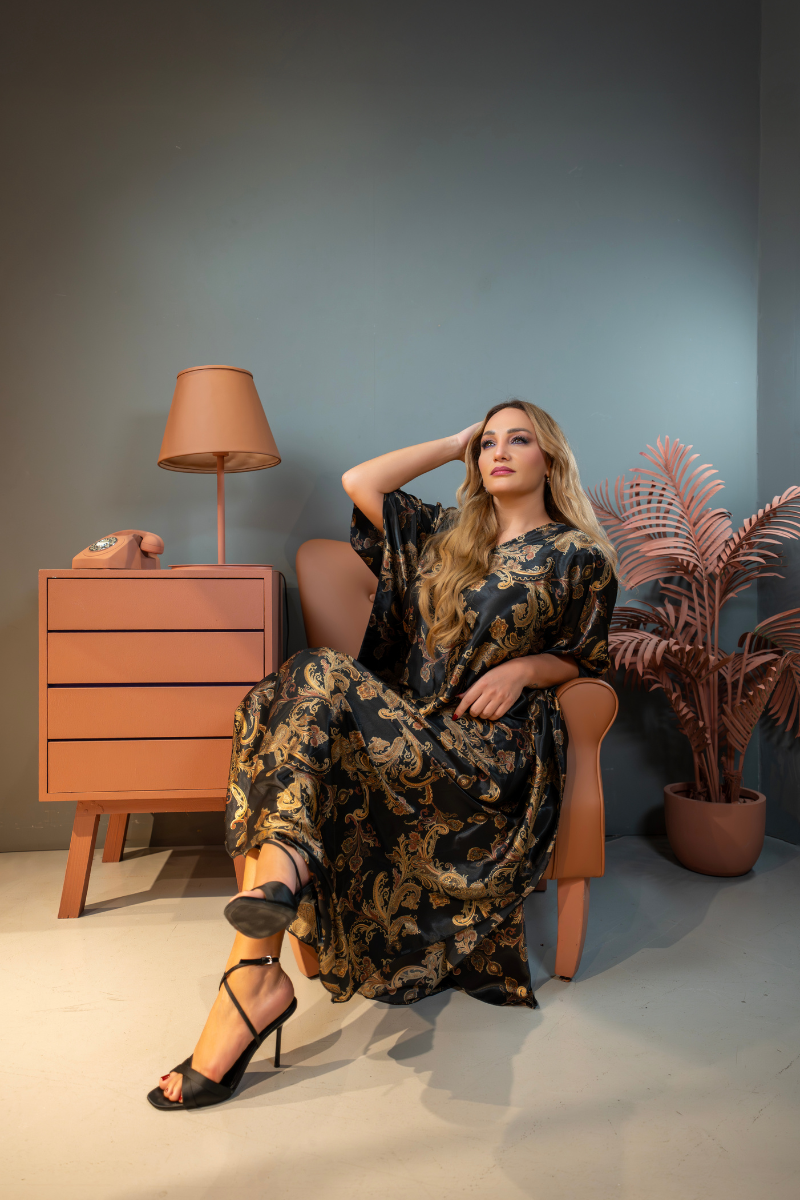 Woman in a patterned dress sitting on a chair next to a lamp and plant against a gray wall.