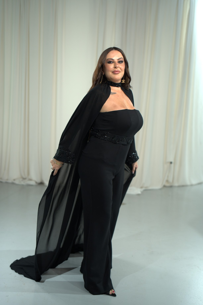 Woman wearing a black outfit with a cape against a white curtain background