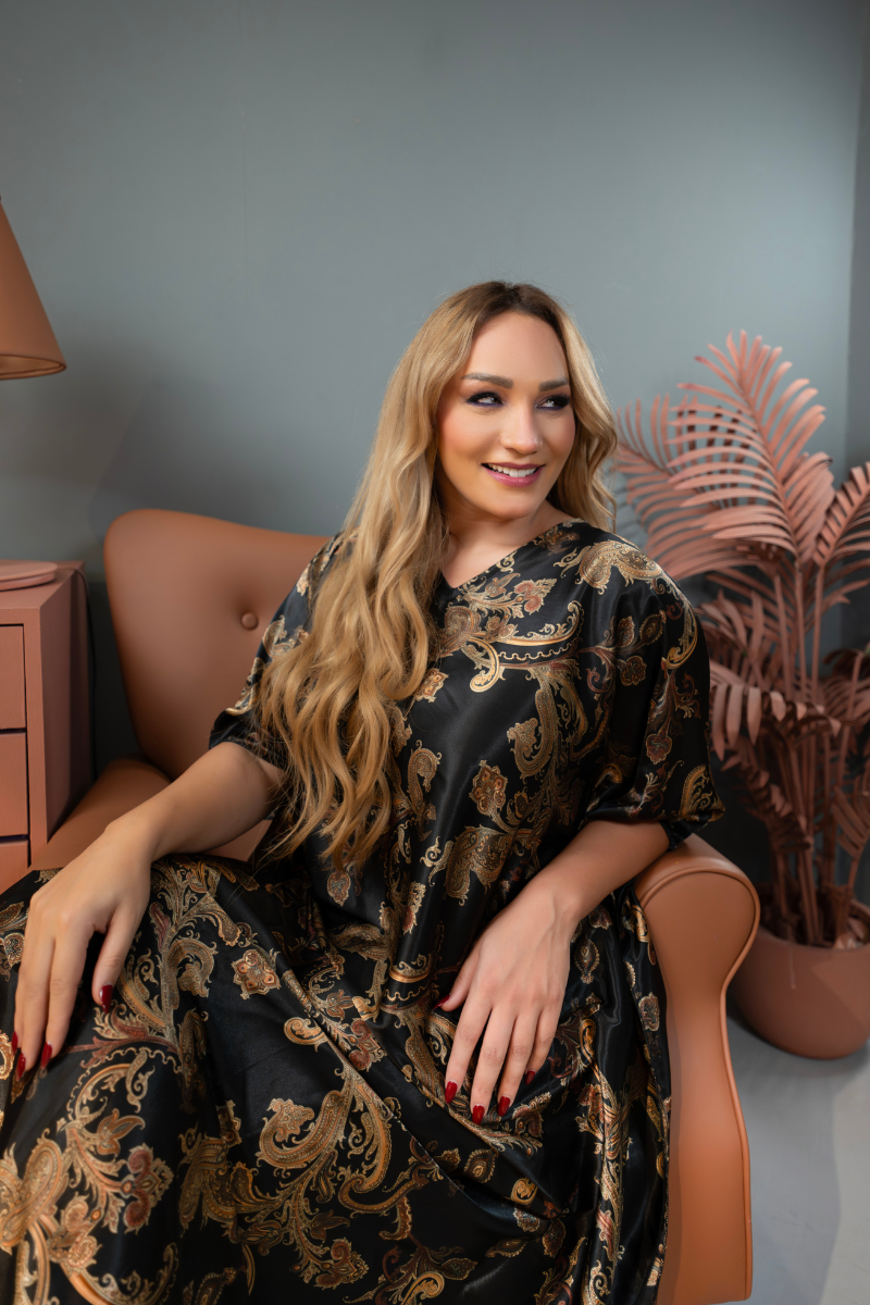Woman in a black and gold patterned dress sitting on a brown chair with a plant in the background.