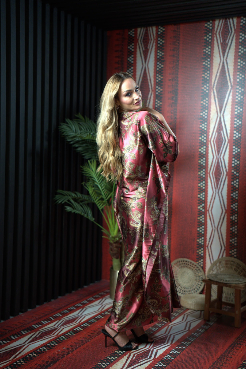 Woman in a patterned dress standing against a decorative wall with a plant and chair.