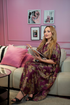 Woman in a patterned dress sitting on a couch with pink walls and decorative frames.