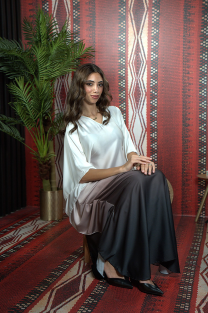 Woman in a white top and dark skirt sitting on a red patterned rug with a plant and wall decor in the background.