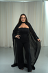 Woman wearing a black outfit with a flowing cape against a white curtain background